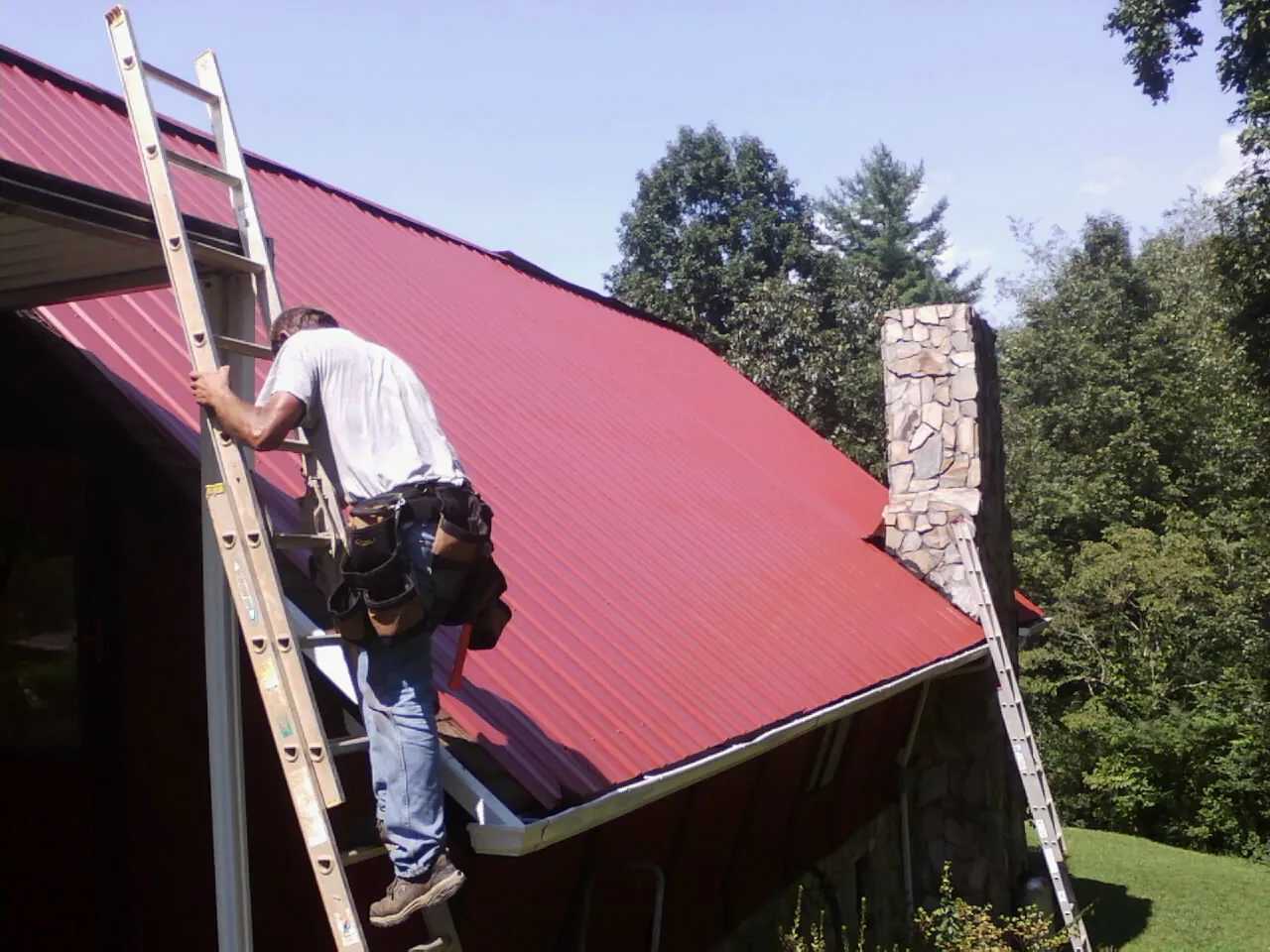 Roofer on a ladder beside a red metal roof and stone chimney — Soffit & Fascia Repair by Roofing Brothers in Stone Harbor